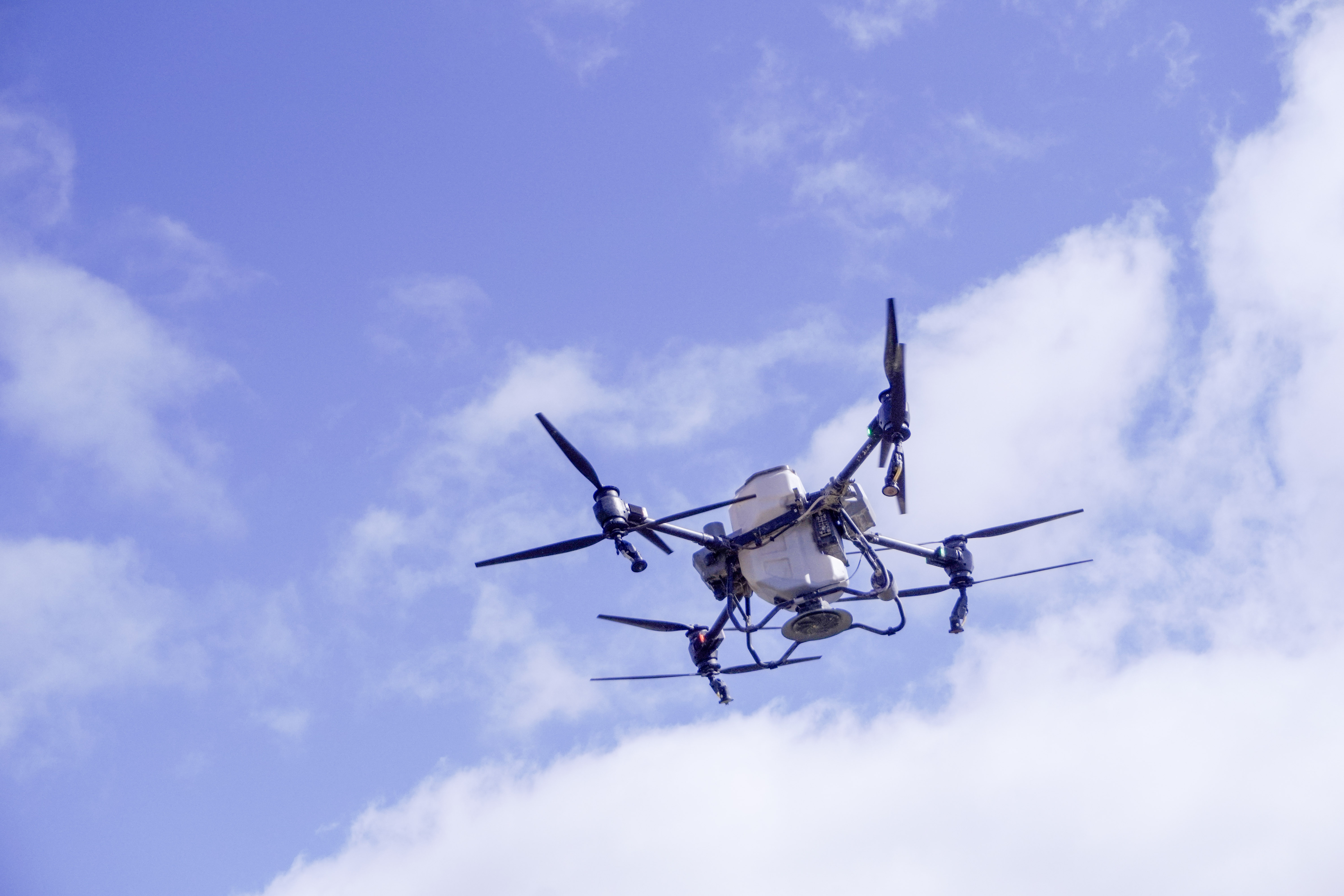 The T50 in flight against a bright blue sky, showing the spreader hopper and spinning disk from below